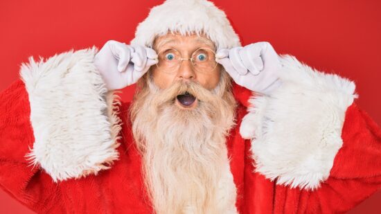 Old senior man with grey hair and long beard wearing santa claus costume holding glasses afraid and shocked with surprise and amazed expression, fear and excited face.
