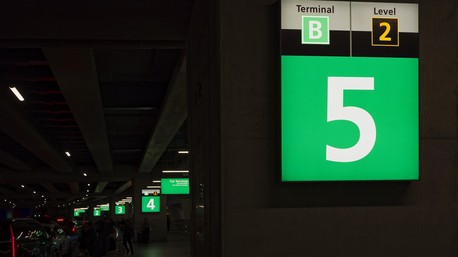 Airport Terminal B Level 2 Gate 5 Sign - A green and white sign with the number 5 on it, indicating the location of a gate in an airport terminal. The sign is mounted on a wall in the airport, with a dimly lit corridor in the background. People are walking in the distance, and there are other signs visible in the background, indicating the location of other gates.